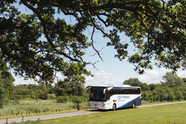 National express bus travelling in the countryside with overhanging branches and a field in the foreground.