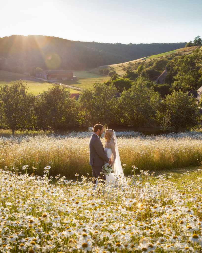 A newly married couple stand looking at each other in a field in the evening sun surrounded by daisies and trees with the hills of the South Downs beyond.