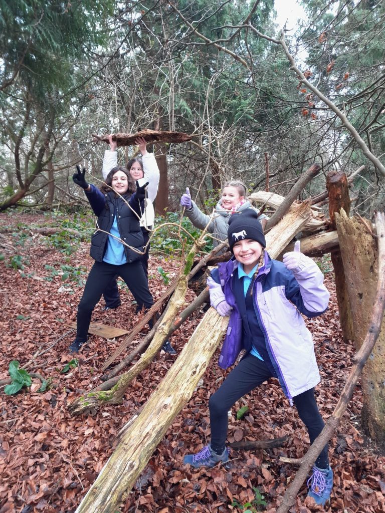 image show primary school children in a woodland setting making a den with branches and giving a thumbs up to the camera