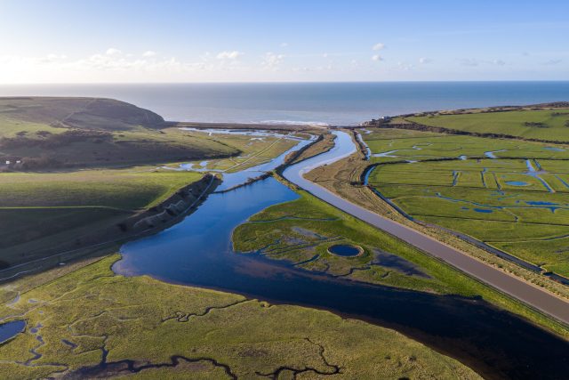 image is a drone shot of the cuckmere meanders at Seven sisters with the sun reflecting in the water and the sea beyond