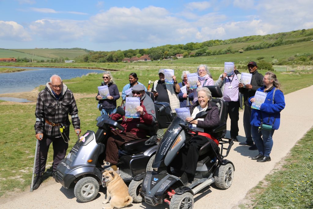 Group of older adults on a sunny countryside outing by a river, smiling and holding "Southdowns View 2026" leaflets, several seated on mobility scooters and a small dog sitting at their feet.