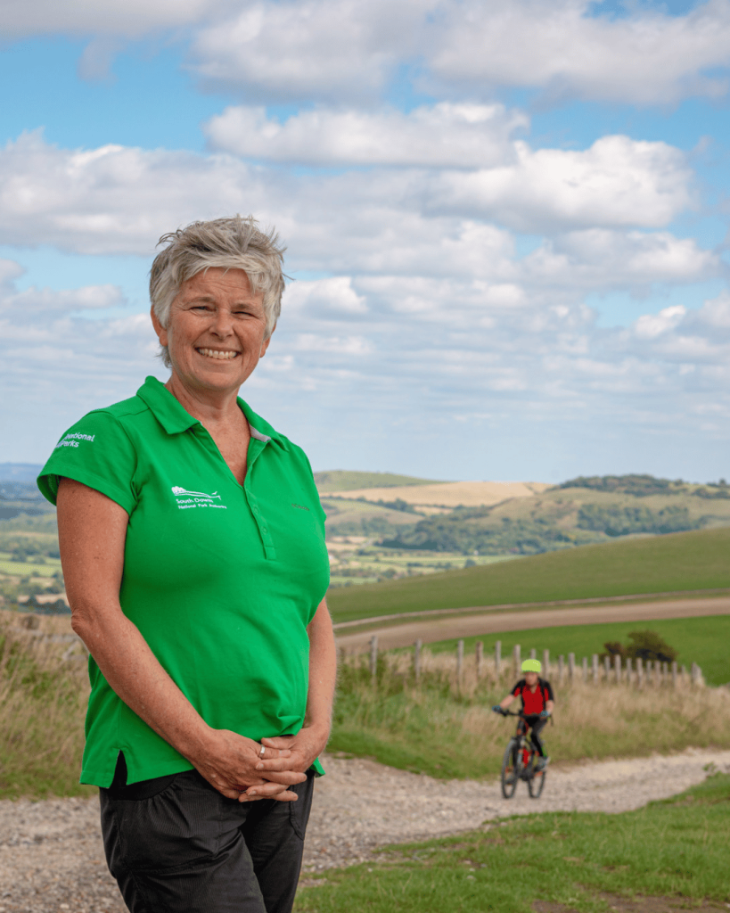 image shows Kate Drake, with a green South Downs National Park Authority t-shirt standing by a path on the south Downs with a cyclist coming up the hill behind her.