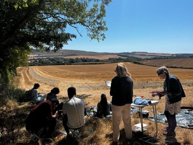 Group of people doing a plein-air painting session on a shaded hillside, some seated sketching or painting while two stand at a small table with art supplies, overlooking golden harvested fields and rolling hills under a clear blue sky.
